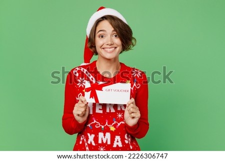 Merry young woman wear knitted xmas sweater Santa hat posing hold gift certificate coupon voucher card for store isolated on plain pastel light green background. Happy New Year 2023 holiday concept