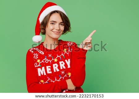 Merry young woman wear knitted xmas sweater Santa hat posing pointing index finger aside indicate on workspace area isolated on plain pastel light green background. Happy New Year 2023 holiday concept
