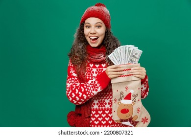 Merry young woman wear red warm cozy knitted sweater hat posing hold Christmas sock fan of cash money in dollar banknotes isolated on plain dark green background. Happy New Year 2023 holiday concept