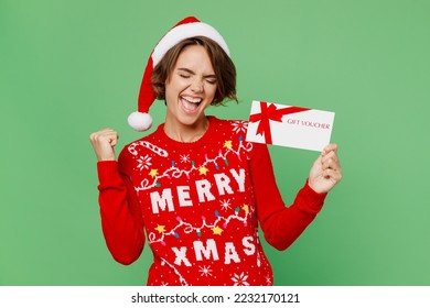 Merry young woman wear xmas sweater Santa hat posing hold store gift certificate coupon voucher card do winner gesture isolated on plain pastel green background. Happy New Year 2023 holiday concept
