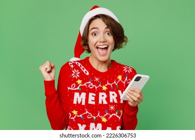 Merry young woman wears knitted xmas sweater Santa hat posing hold in hand use mobile cell phone do winner gesture isolated on plain pastel light green background. Happy New Year 2023 holiday concept