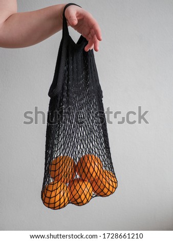 Mesh bag with fruits in female hand. Hand of Stylish young woman hold mesh shopping bag on light gray wall. Modern reusable shopping, zero waste concept. Eco frendly