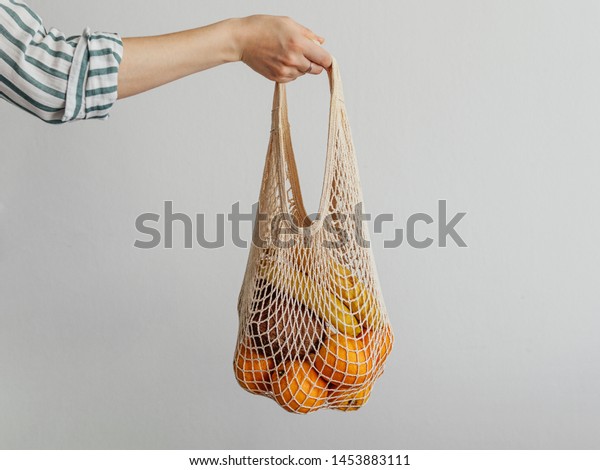 Mesh bag with fruits in female hand.\
Hand of Stylish young woman hold mesh shopping bag on light gray\
wall. Modern reusable shopping, zero waste\
concept.