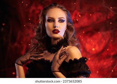 Mesmerizing beautiful witch. An attractive sexy vampire girl in a black dress stands in an old castle in blood red lighting. Halloween. Vampires.  