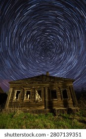 Mesmerizing star trails create concentric circles above a weathered, abandoned house in rural America, its wooden frame illuminated by artificial light against the dark landscape.