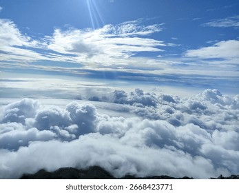 A mesmerizing view from above the clouds, where fluffy white formations stretch across the horizon under a radiant blue sky with beams of sunlight piercing through.

