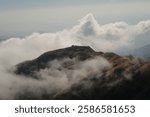 A mesmerizing view of a remote mountain settlement sitting atop a golden-hued ridge, partially enveloped by misty clouds. The contrast between the rugged landscape and the clouds creates a dreamlike