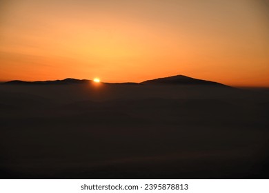 Mesmerizing view of the sun setting behind sharp mountain peaks shrouded in mist. Sky filled with bright colors surrounds winter mountains.
