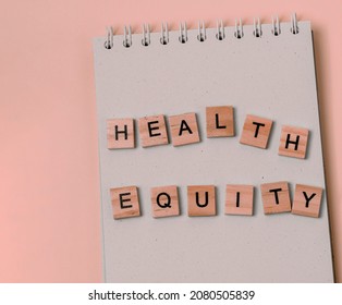 Message Health Equity With Wooden Letters On Notebook In Pink.