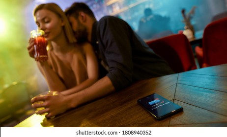 Message from wife. Unfaithful man spending time with another woman in the bar, enjoying drinks and conversation. Cheating, infidelity. Focus on smartphone, wife or girlfriend is texting. Web Banner