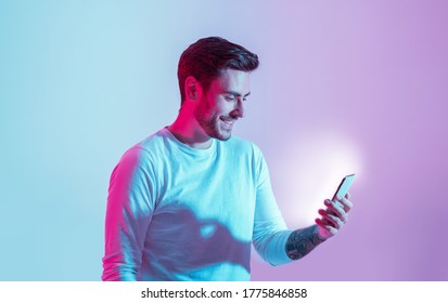 Messages from social network. Smiling young man look at luminous smartphone in neon light, copy space