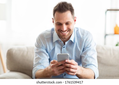 Messaging At Social Network. Happy millennial guy using modern smartphone device, sitting on sofa at home