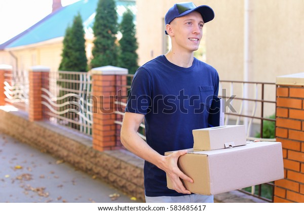 Messenger Carring Two Boxes in Hands Smiling on\
Background of Trees Along Street. Courier Company Provides Its\
Services in All Areas Ofterrain, Man of European Appearance in\
Uniform Carries Two\
Boxes