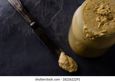 A Messy Jar Of Homemade Peanut Butter In A Glass Jar Sitting On A Dark Gray Slate With An Antique Silver Speading Knife.