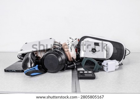 Messy Pile Of Discarded Electronic Devices Including Camera And Headphones Sits On Desk Against Plain Background