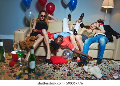 Messy room after wild house party, three tipsy stylish friends relaxing on couch while blond-haired woman with teddy bear posing for photography