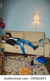 Messy Room After Wild Party: Colorful Confetti Thrown Everywhere, Empty Alcohol Bottles And Champagne Flutes Standing On Floor And Coffee Table, Asian Man Sleeping On Couch