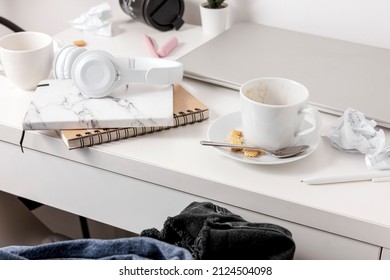 Messy table with laptop, dirty cups, and stationary items. Cluttered workplace, desk. Concept of being overwhelmed by working or studying. Mess and chaos at home. Deadline, stress.