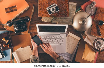 Messy Vintage Desktop With Laptop And Male Hands Using A Smart Phone, Top View