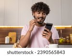 A mestizo guy in the kitchen in subdued lighting, holds a phone and potato fries in hands, eats seductively with pleasure and reads romantic message in the screen. A young american or african man