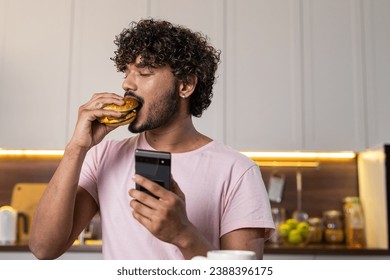 A mestizo guy stands in profile in the kitchen in subdued lighting, holds a phone and hamburger in hands, seductively eats a burger with closed eyes in pleasure. A young american or african man enjoys