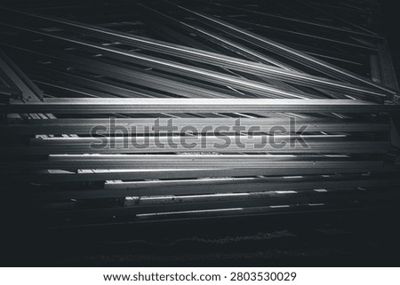 Metal beams stack darkly, gleaming under dim light. Rows of steel intersect with sharp angles and shadowed depth. Industrial decay feels heavy, yet orderly in its disarray. Ideal for construction