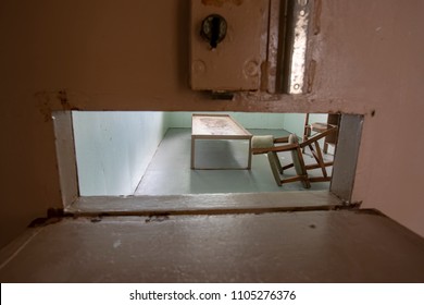 Metal Bed Frame With Chair And Desk Inside Solitary Confinement Cell Through Metal Door Slat In Old Prison.