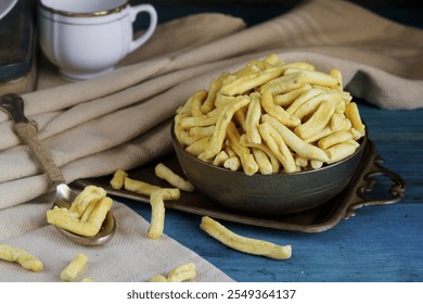 A metal bowl brimming with crunchy, yellow snacks is displayed on a vintage tray, accompanied by a spoon, against a blue wooden background.