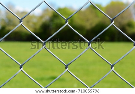 Metal chain link fencing in selective focus. Beyond the secure fence lies an empty green field, lined by trees.
