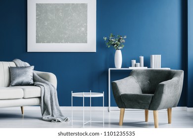 Metal coffee table, grey armchair, white sofa with a blanket and pillow, and big painting on the blue wall in a living room interior - Shutterstock ID 1101235535