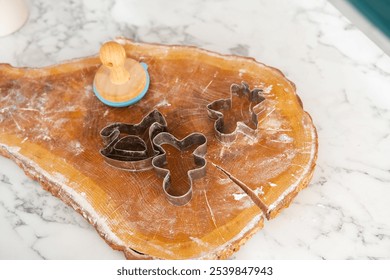 Metal cookie cutters shaped like holiday figures lie on a wooden cutting board dusted with flour. A stamp rests nearby, ready for festive baking in a sunny kitchen. - Powered by Shutterstock