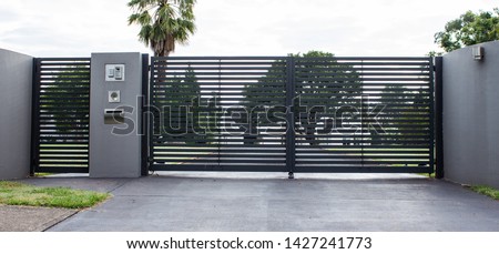 Metal driveway property entrance gates set in concrete fence with garden trees  in background