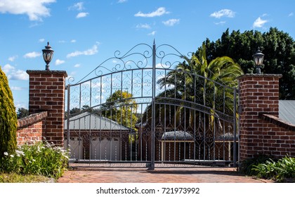 Metal driveway security entrance gates set in brick fence with residential garden in background against blue sky