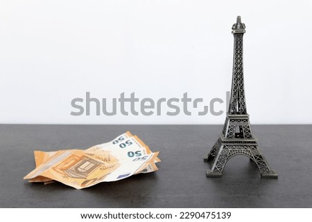 Metal Eiffel Tower statue and fan of 50 euro bills on black table surface against white wall background, conveying a tone of luxury and wealth associated with visiting Paris.