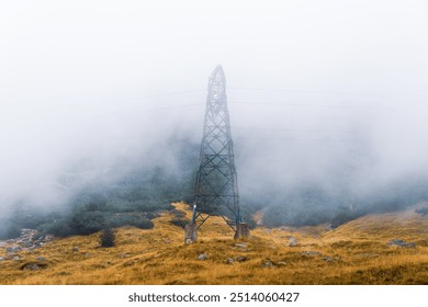 A metal electricity tower stands in a foggy mountain landscape. Yellow grass and green bushes on the slopes contrast with the thick mist covering the sky, creating a serene, moody atmosphere. - Powered by Shutterstock