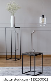 Metal End Table Standing On Carpet In Real Photo Of Bright Room Interior With Glass Lamp, Wainscoting On Wall And Flowers In Vase Placed On Stand In Blurred Background