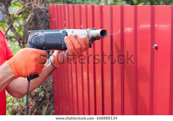 Metal fence installation. Work man in gloves\
with a drill builds new metal\
fence.