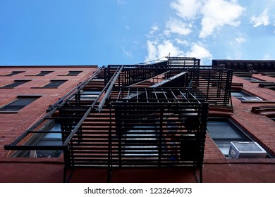 Metal Fire Exit Stairs On The Exterior Brick Wall Of A City Apartment Building. A Few Clouds In A Blue Sky Above. Bottom Looking Up View Point. Greenpoint, Brooklyn, New York, NY.         