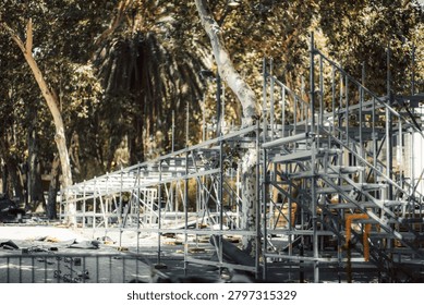 Metal framework of a temporary stage being installed or dismantled among trees in a public park, with scaffold stairs, event structure and scattered equipment