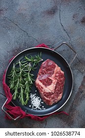Metal frying pan with fresh uncooked grass-fed beef steak, rosemary and salt. Flat-lay with copyspace on a weathered asphalt background