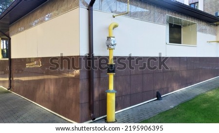 A metal gas pipe with a shut-off and safety valve exits the ground and enters a building lined with ceramic tiles. there is a drain pipe nearby. The blind area of the building is made of concrete tile