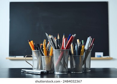 metal glass with pens, pencils and felt-tip pens and scissors on the background of an empty black chalk board