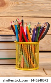 Metal holder with multicolored pencils. Stack of books and colorful pencils in basket on wooden background.