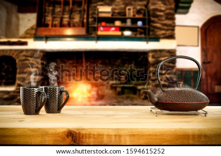 Metal kettle of hot drink.Wooden board with empty space for your products.Blurred background of retro fireplace and orange light of fire.Hot tea, coffee or mulled.Cold winter night in home interior. 