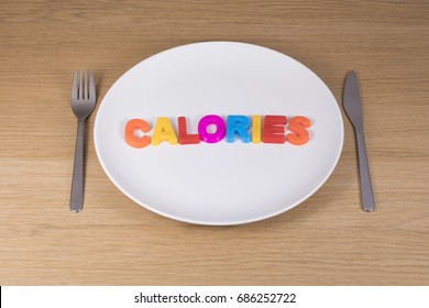 A Metal Knife, Fork And Empty White Plate With The Word Calories Written In Colourful Toy Learning Letters Against A Plain Timber Bench Top Background.