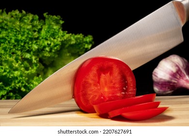 A metal knife slicing a red ripe tomato on a wooden board. Close up of a sharp knife blade cutting tomato on slices, green salad and garlic on a black isolated background. - Shutterstock ID 2207627691