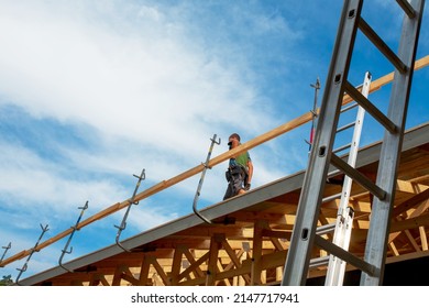 Metal Ladder In Front Of Wooden House Under Construction Whith Young Male Worker Walking On The Roof On A Sunny Summer Day