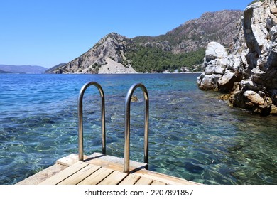 Metal ladder to the sea on wooden pier, picturesque view to mountains and bay with transparent water. Beach vacation and swimming concept