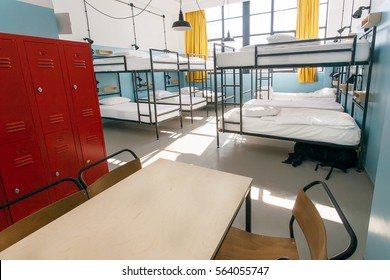 Metal locker, table and double-decker beds inside hostel room with tall windows.
