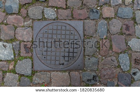 Metal manhole cover. The road is paved with granite cobblestones. Old construction on the sidewalk.